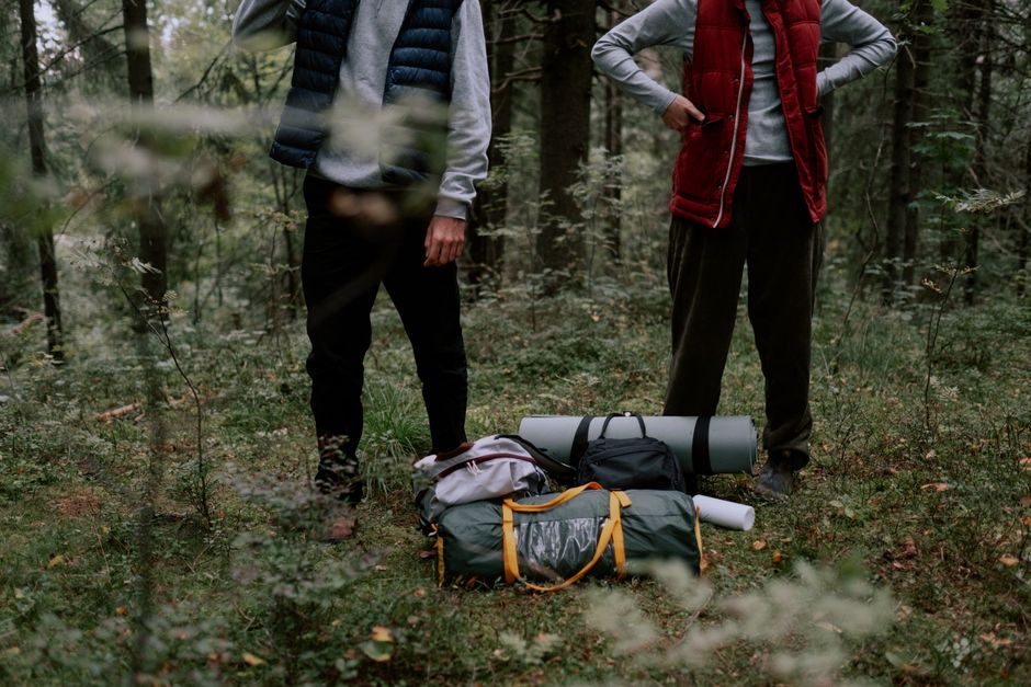 Two adults standing with camping gear in a forest, surrounded by greenery.